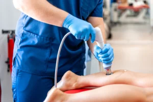 A person in blue scrubs and gloves uses a handheld ultrasound device on someone's leg. The setting appears clinical, focusing on the treatment process.