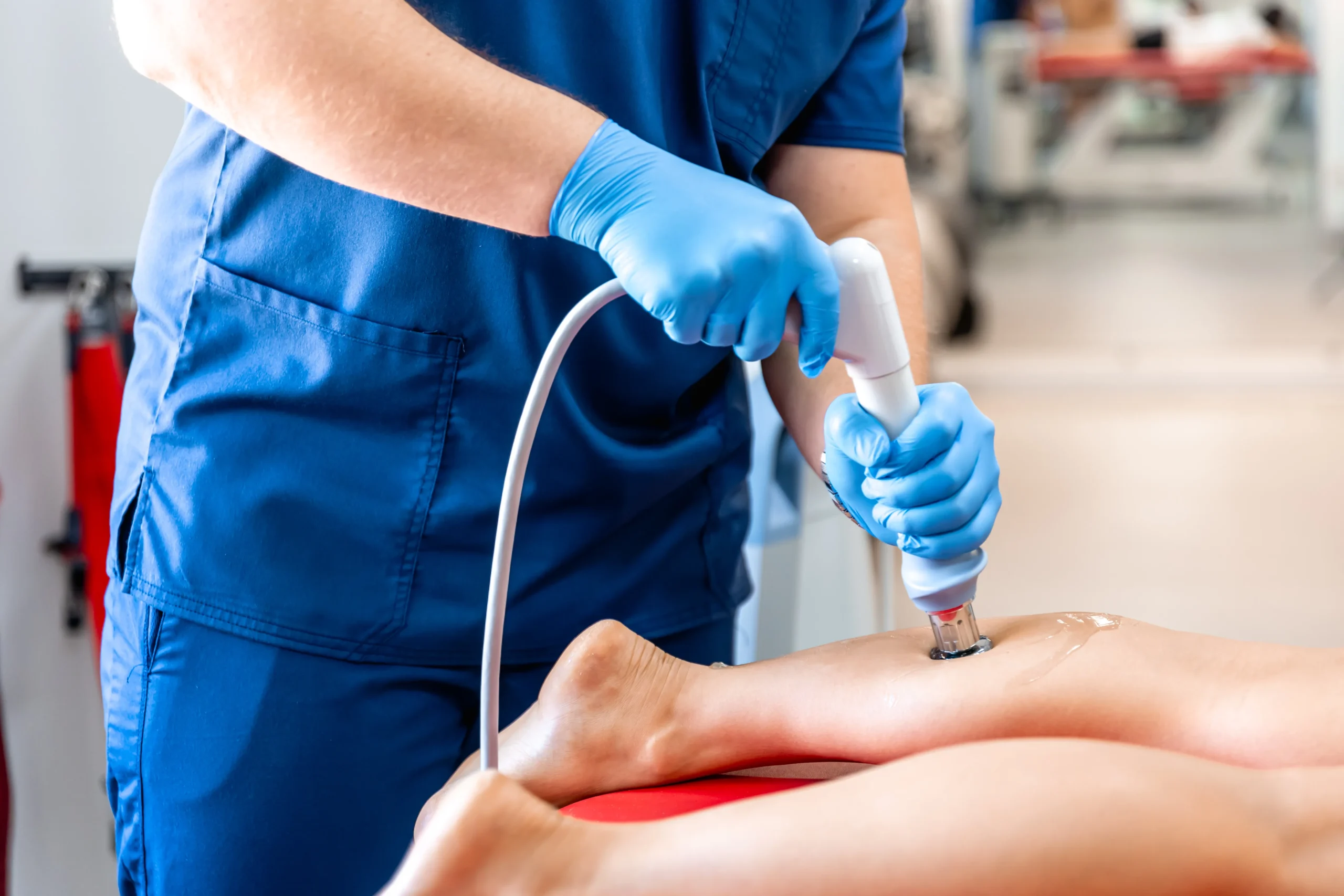 A person in blue scrubs and gloves uses a handheld ultrasound device on someone's leg. The setting appears clinical, focusing on the treatment process.