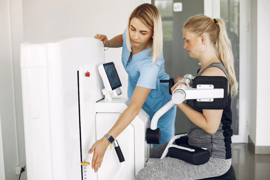 A physiotherapist in blue scrubs adjusts a seated exercise machine for a woman in activewear. The setting appears clinical and supportive.