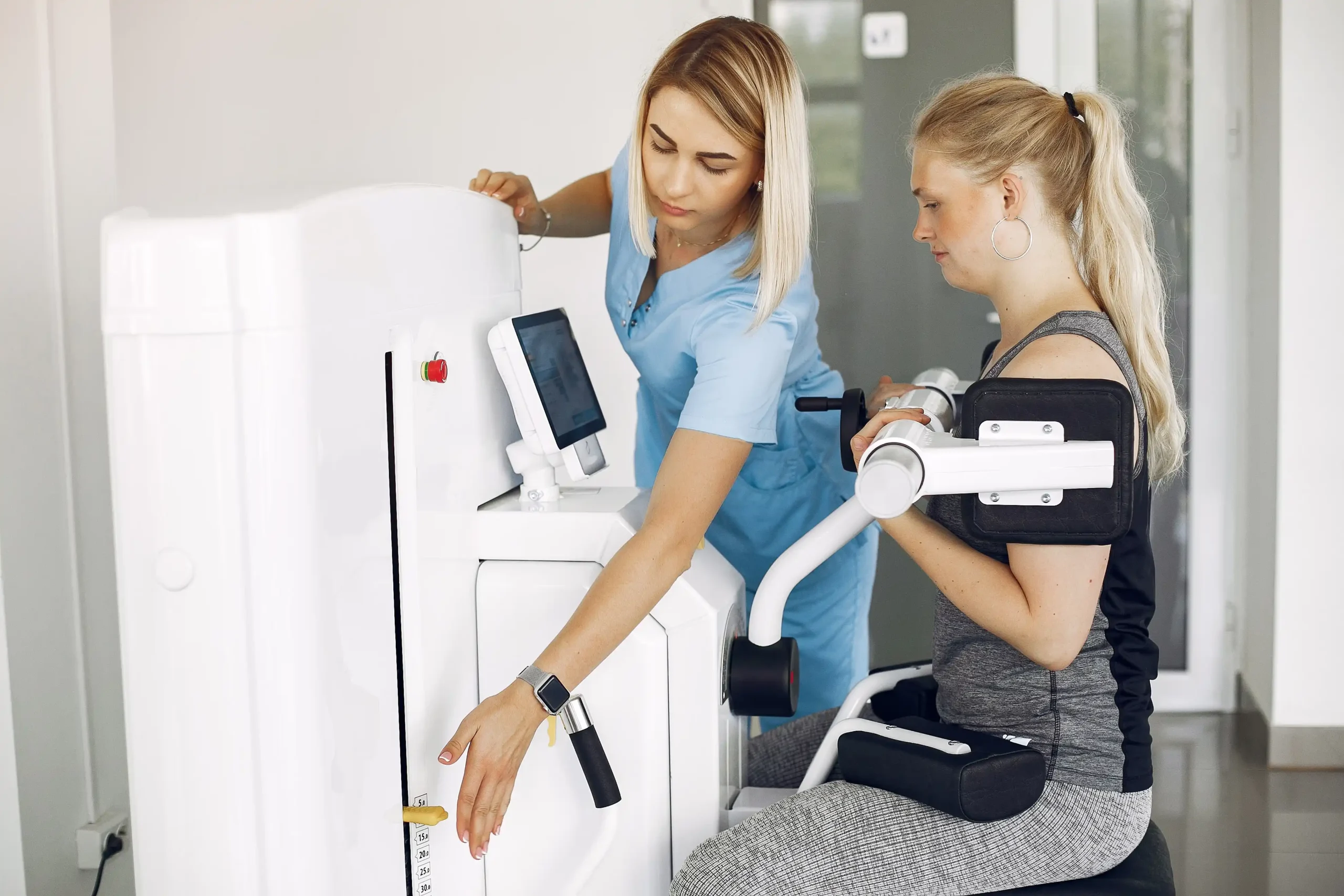 A physiotherapist in blue scrubs adjusts a seated exercise machine for a woman in activewear. The setting appears clinical and supportive.