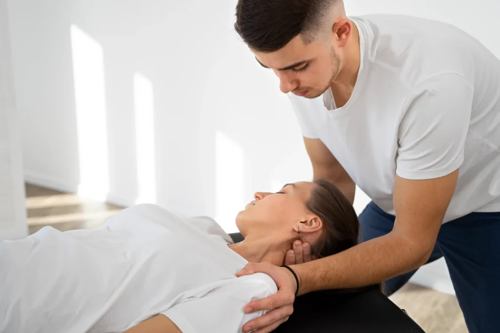 A therapist in a white shirt performs neck adjustments on a relaxed woman lying on a massage table in a bright room, conveying calm and care.