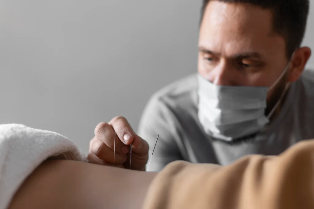 A practitioner wearing a mask focuses intently while inserting acupuncture needles into a person's back, conveying a sense of calm and precision.