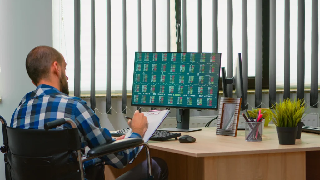A man in a wheelchair, wearing a blue plaid shirt, reviews notes at a desk with a computer displaying financial data. The setting is an office with vertical blinds, plants, and office supplies, conveying focus and productivity.