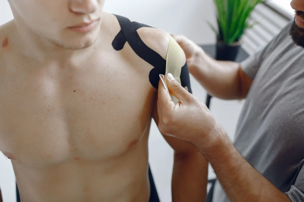 Person applying black kinesiology tape to a shirtless man's shoulder. The setting is a bright room with a potted plant, conveying a clinical tone.