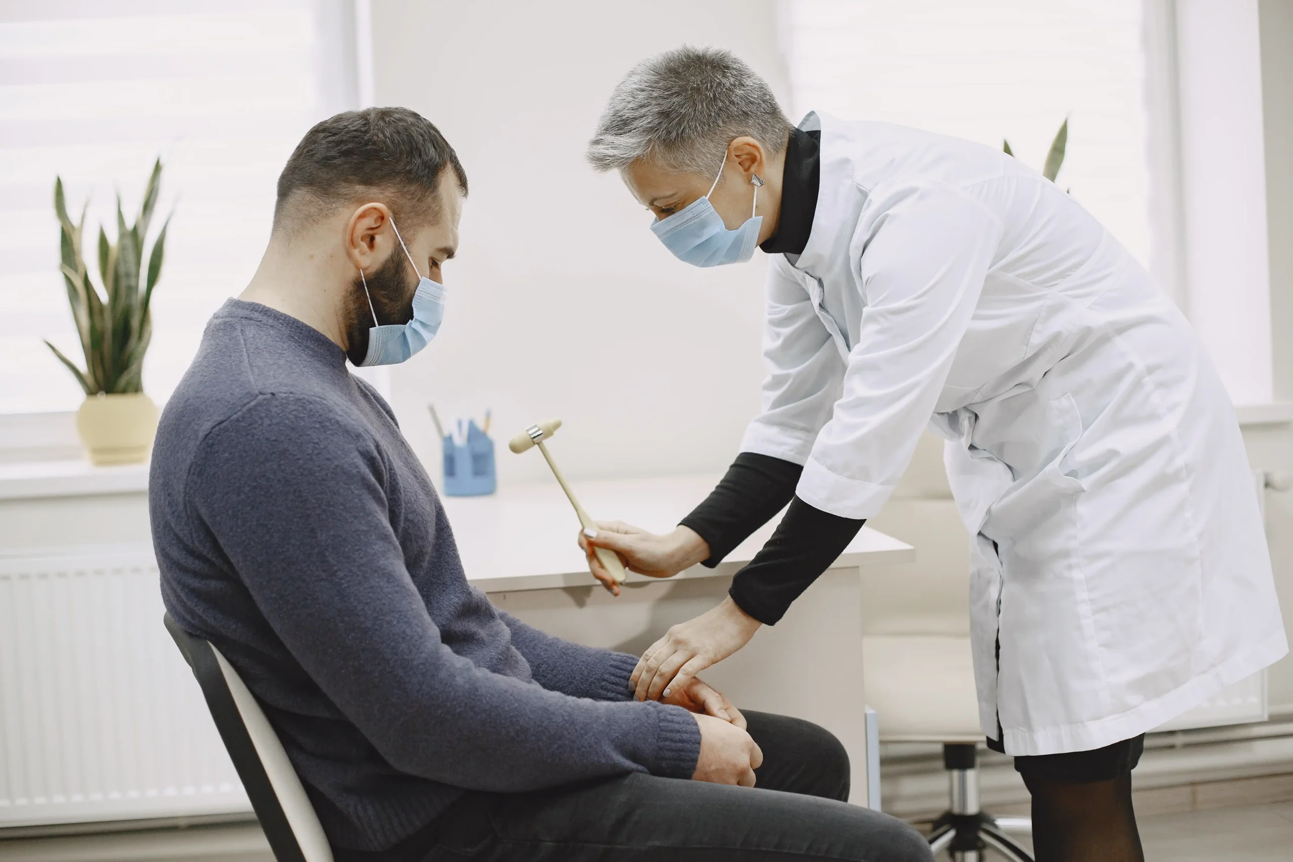 A healthcare professional in a white coat performs a knee reflex test on a seated male patient. Both wear masks, emphasizing a clinical setting.