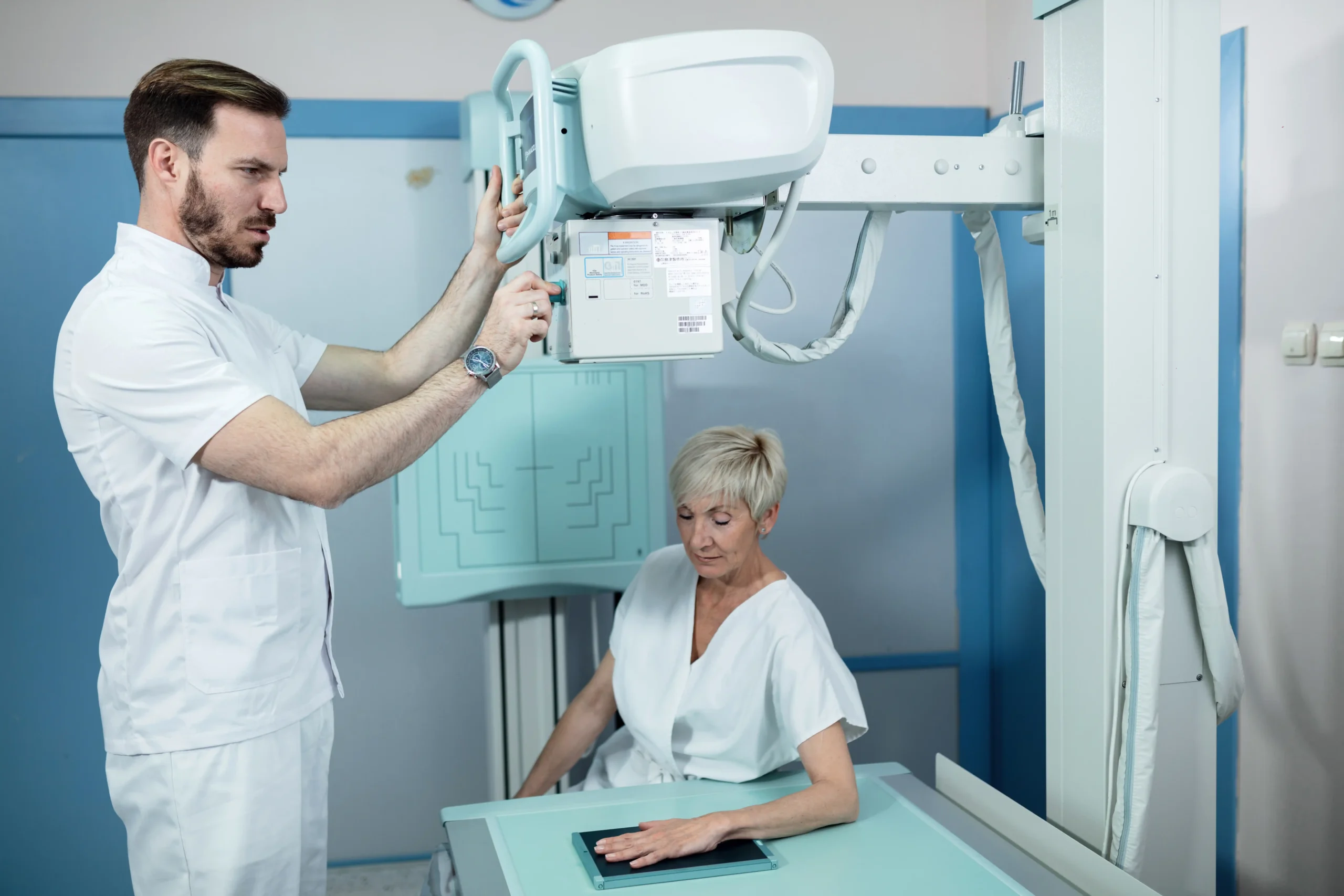A healthcare professional adjusts an X-ray machine above a seated, older woman placing her hand on the table. The setting is clinical and focused.