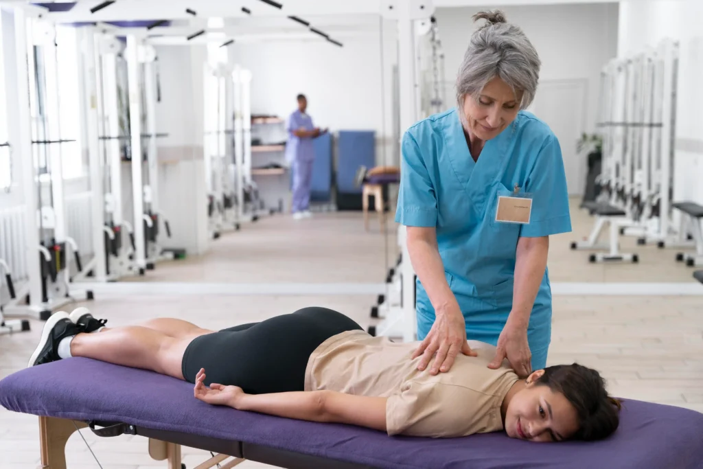 A female physiotherapist in blue scrubs massages the back of a woman lying face down on a purple table in a bright, spacious clinic.