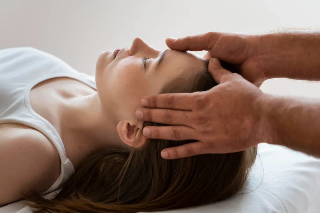 A woman lies calmly on a massage table, eyes closed, receiving a gentle head massage from a pair of hands, creating a serene and relaxing atmosphere.