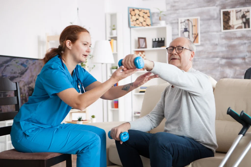 A nurse assists an elderly man in arm exercises with dumbbells in a cozy living room. The scene conveys support and encouragement in a home care setting.