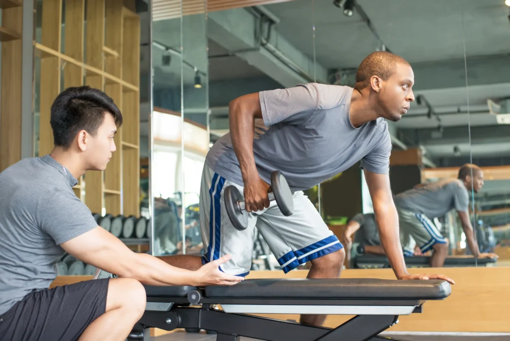 A man in a grey shirt performs a dumbbell row on a bench in a gym. Another man kneels beside him, offering guidance. The setting is focused and supportive.