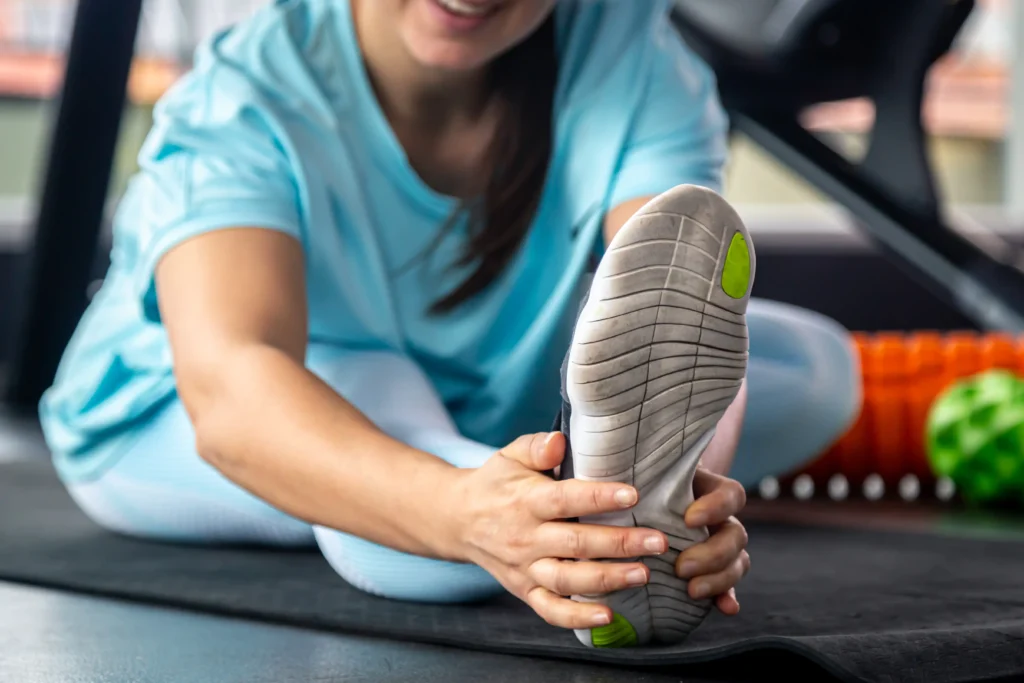 A woman in athletic wear stretches her leg, smiling on a gym mat. Nearby, orange and green foam rollers suggest a fitness setting.