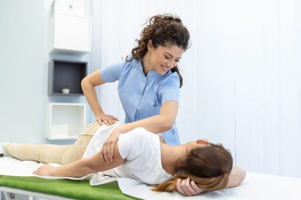 A smiling physical therapist in blue scrubs performs a stretch on a patient lying on a table in a bright, minimalist clinic. The scene conveys care and relaxation.