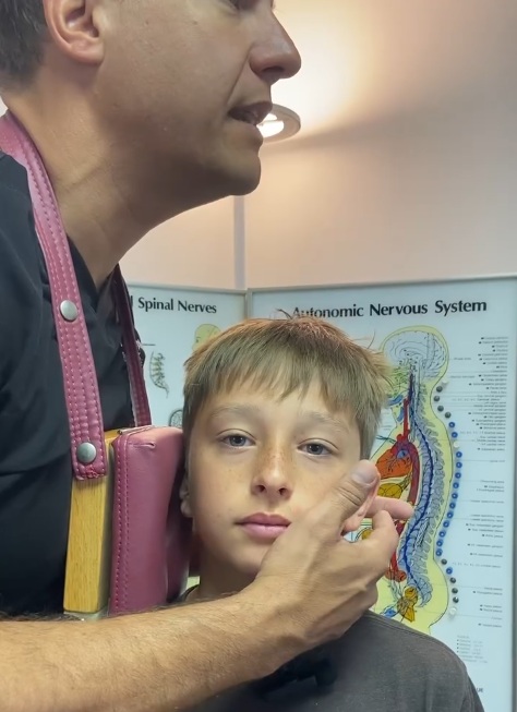 Pediatric chiropractor performing a gentle spinal adjustment on a child at a New York, NY clinic with an autonomic nervous system chart in the background.